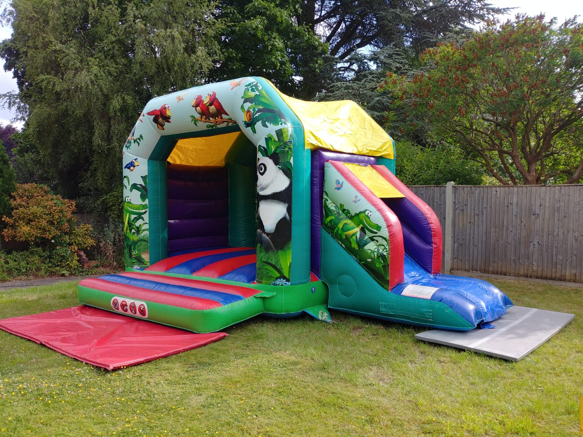 A Majestic, green, blue, red, and yellow, inflatable castle for hire from 121 Bounce with a design of birds, reptiles, mammals, and insects in a wilderness setting. There are also elements typical of a playground, such as trees and plants, adding to the fun of this park-like setting.
